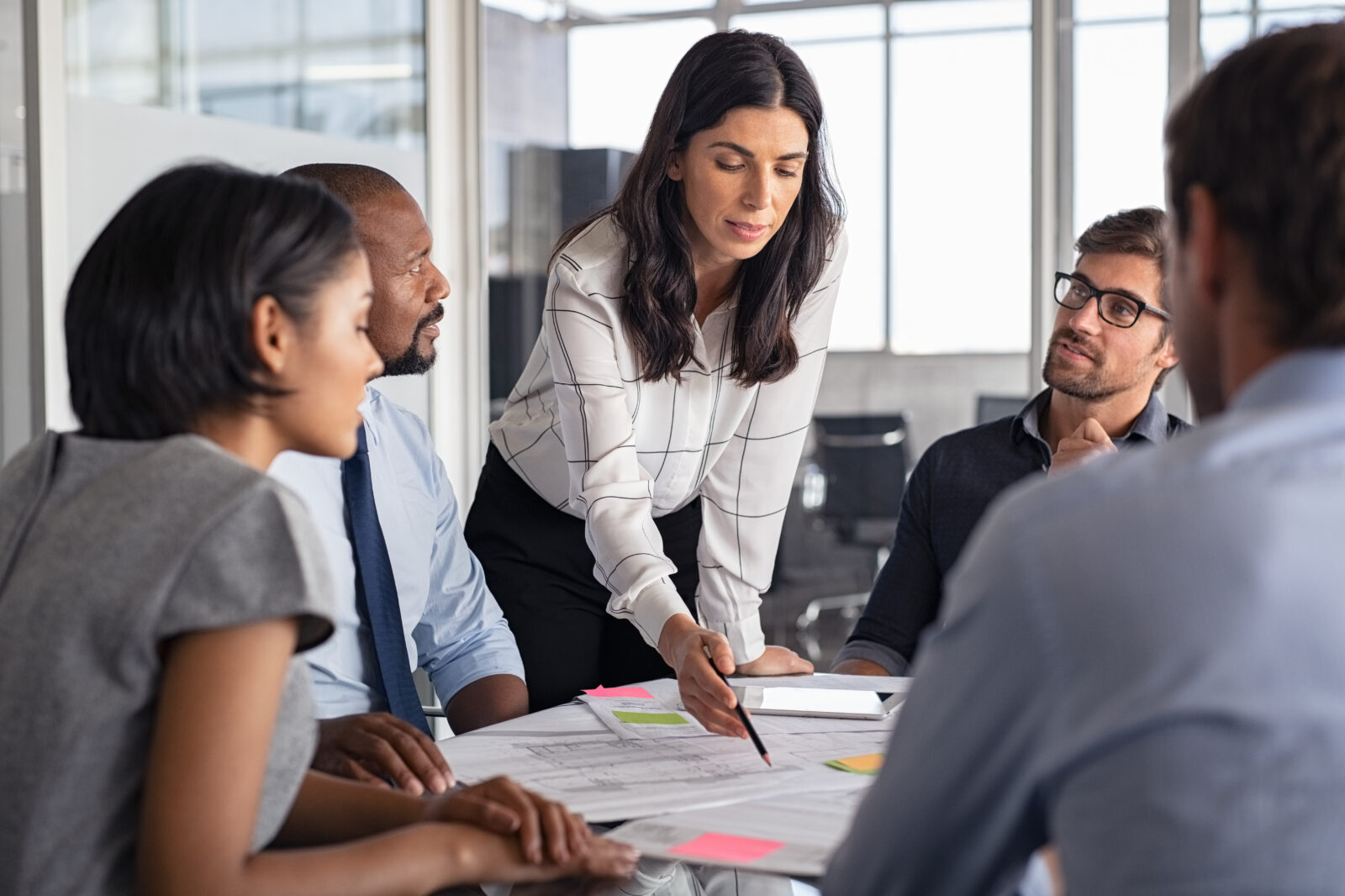 Woman with long brown hair standing over table of colleagues pointing at and presenting paperwork in a business discussion.